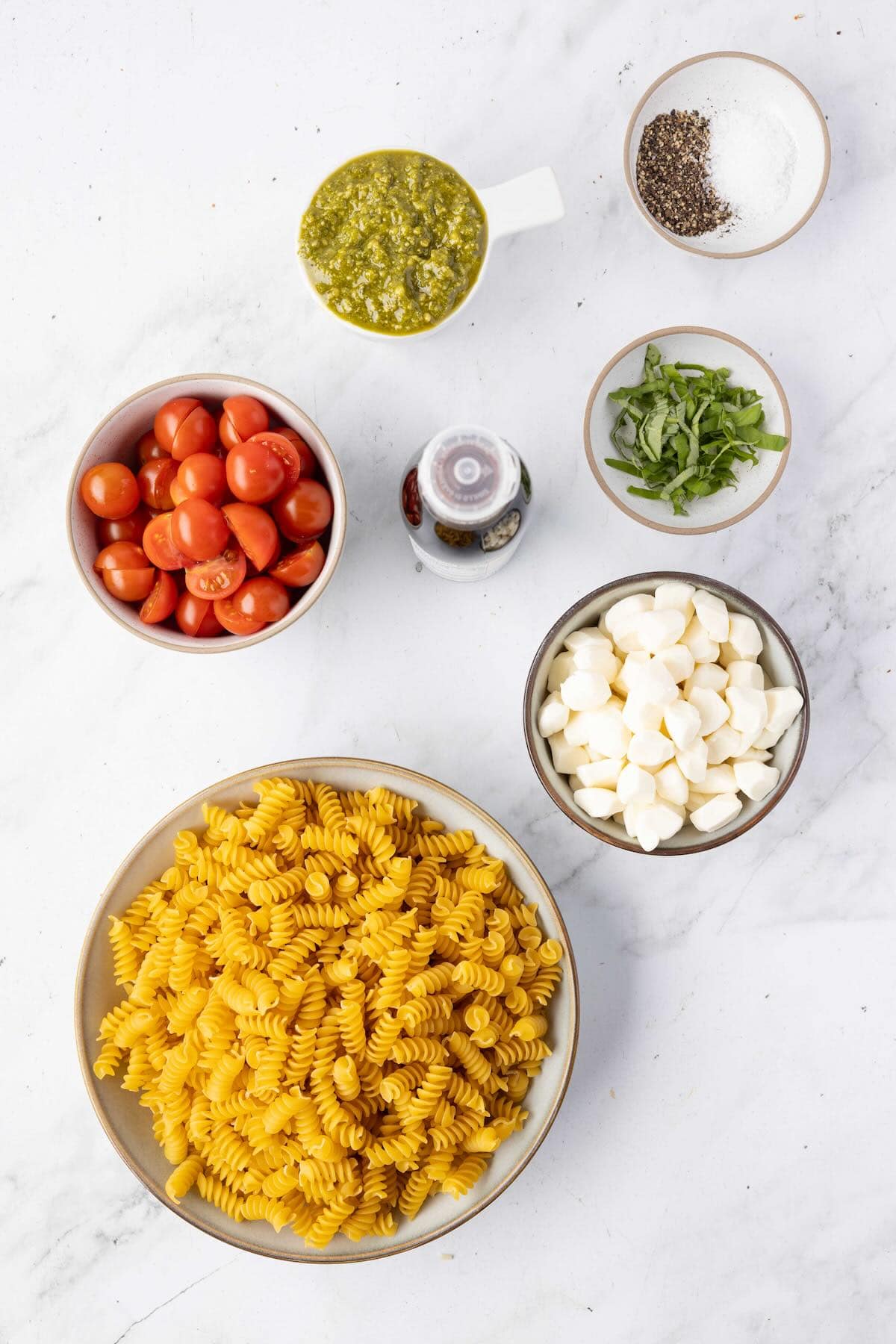 Ingredients for Caprese pasta salad including cherry tomatoes, mozzarella pearls, pesto, basil, and rotini pasta arranged on a kitchen counter.