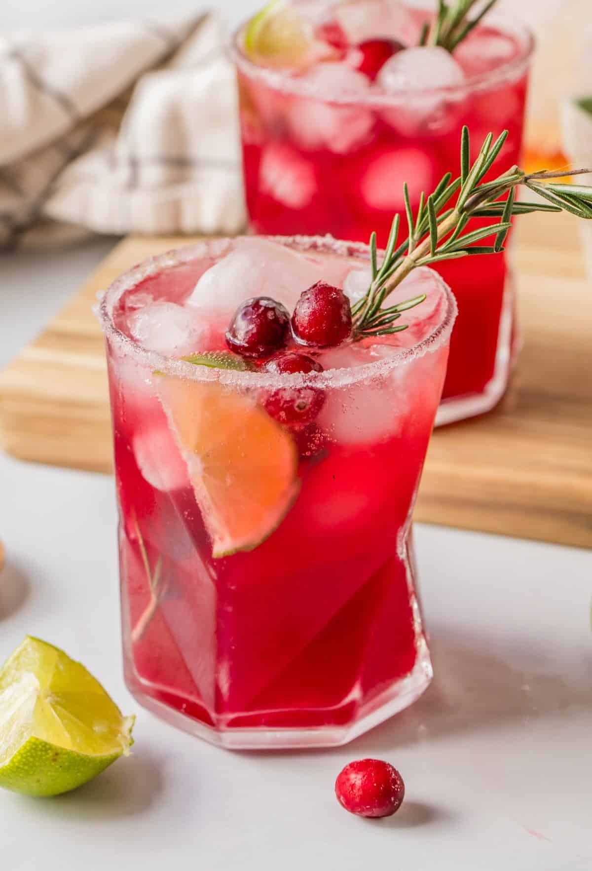 Two cranberry lime margarita mocktails garnished with rosemary sprigs and lime wedges for a festive look.