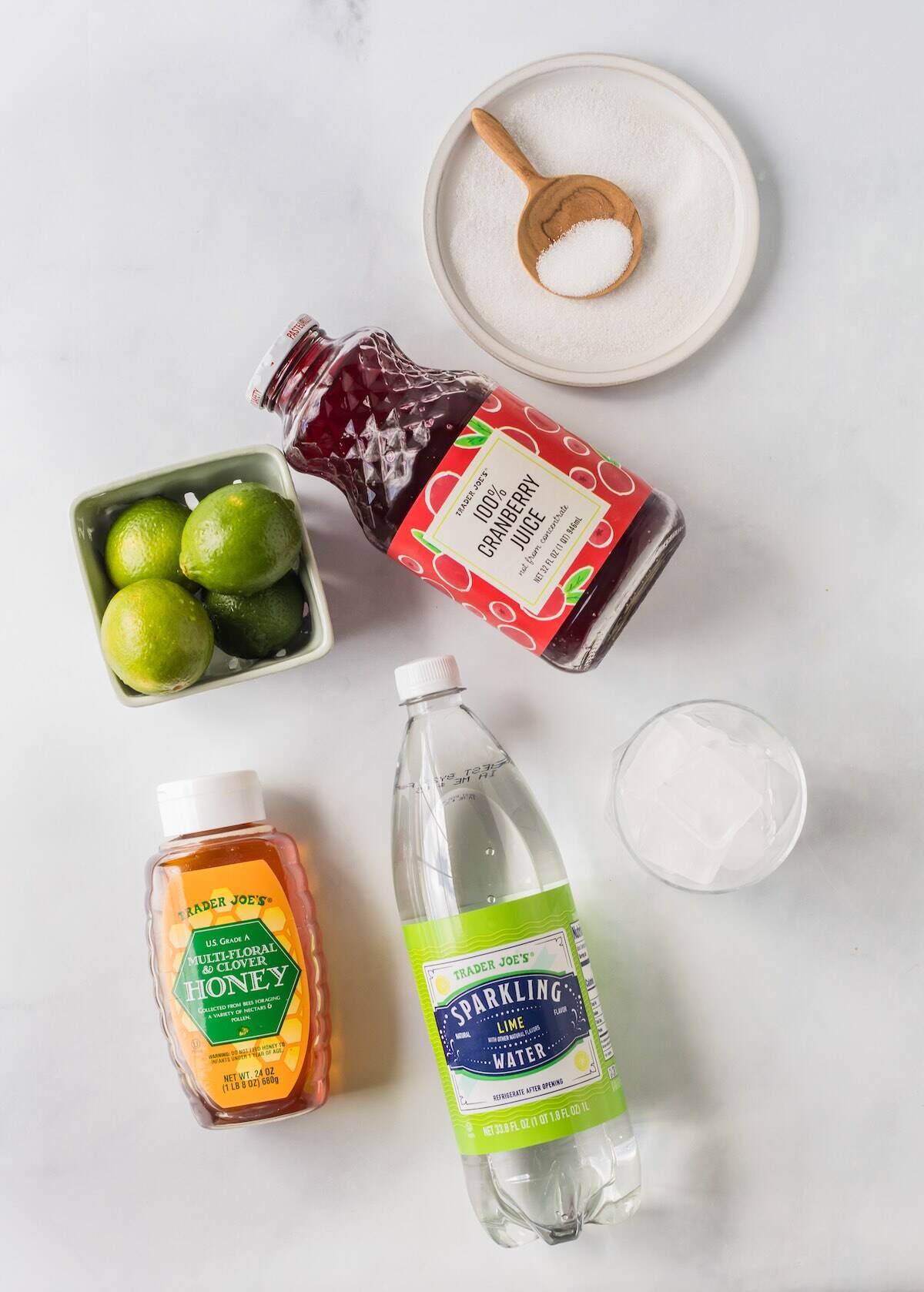 Overhead view of ingredients for cranberry lime margarita mocktail, including cranberry juice, limes, honey, and sparkling water