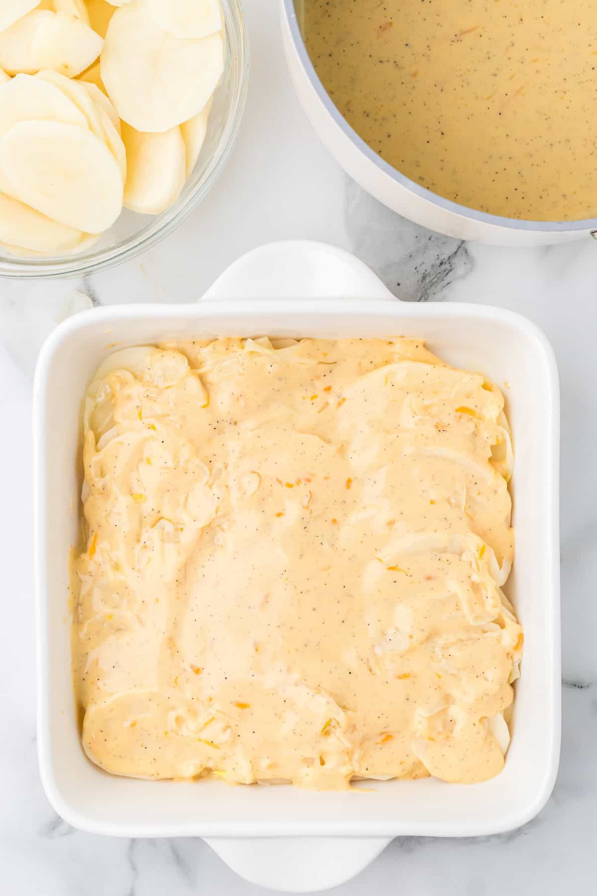 First layer of sliced potatoes and onions in a casserole dish topped with cheese sauce.
