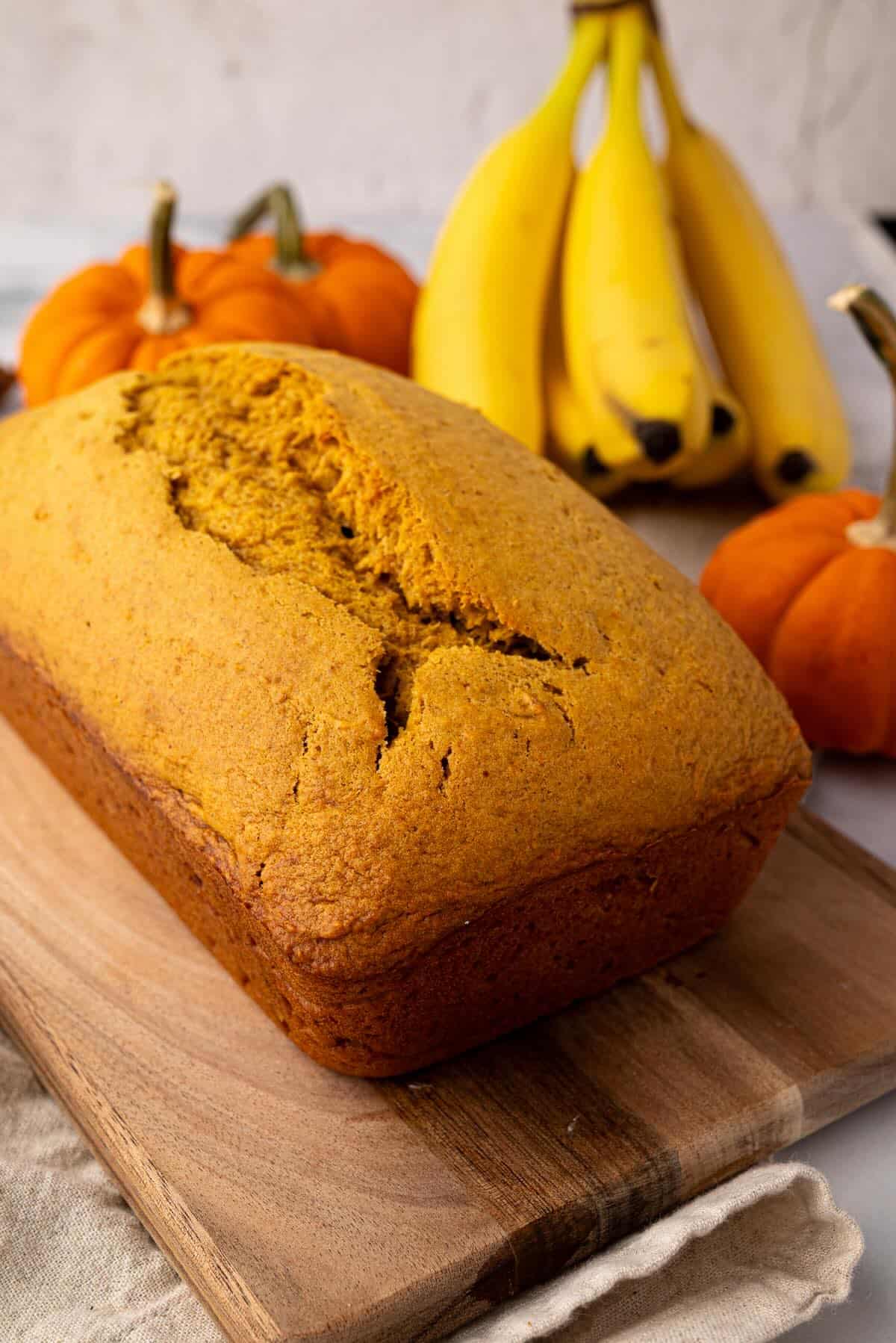 Freshly baked pumpkin banana bread loaf on a cutting board rack with pumpkins and bananas in the background.