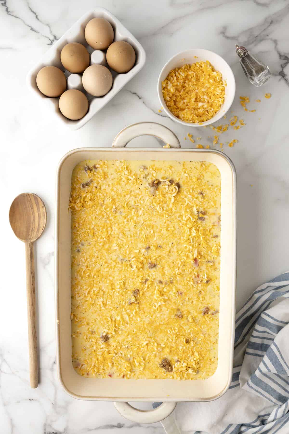 Unbaked egg casserole sprinkled with shredded cheese in a baking dish.