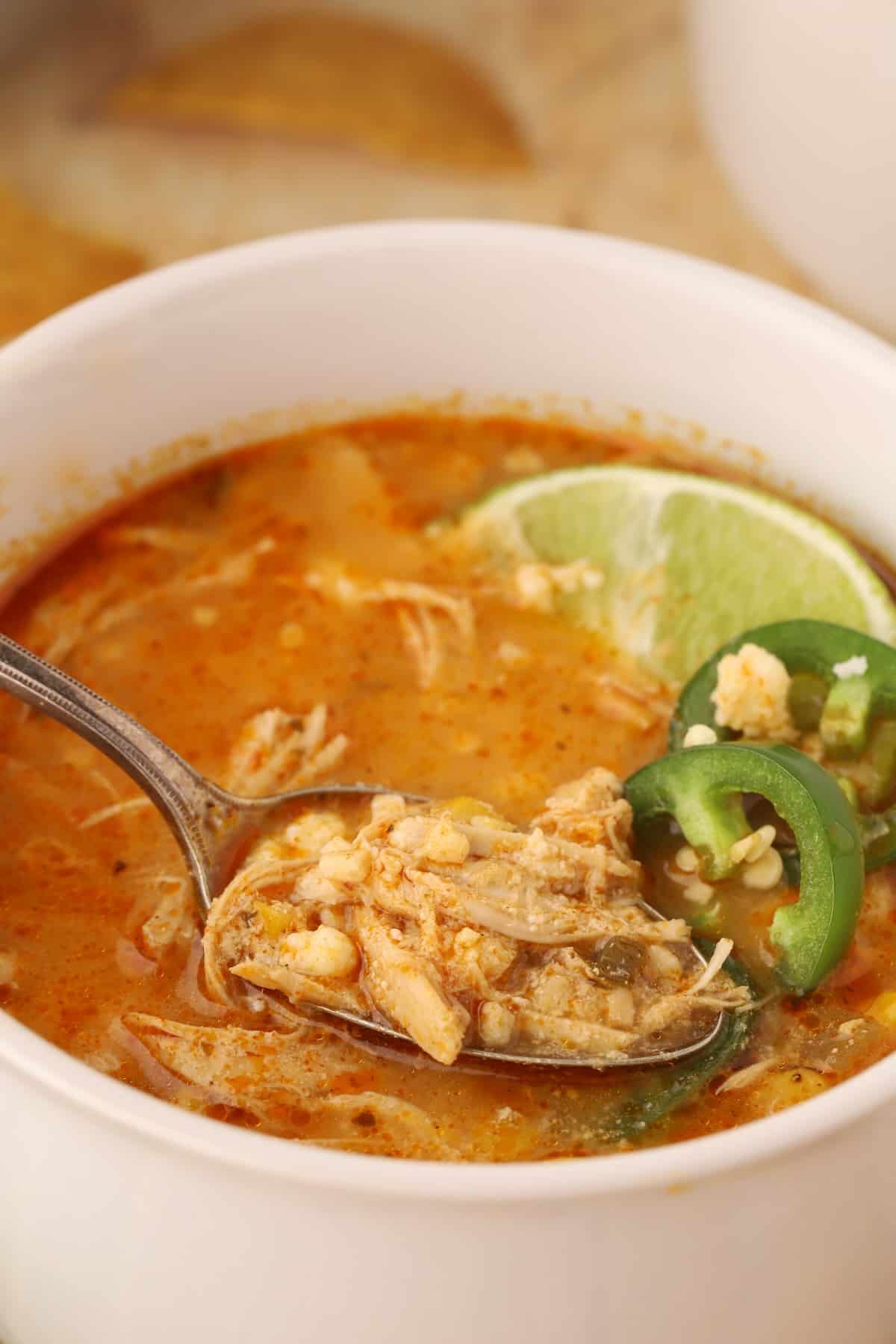 A spoonful of soup showing a rich broth, tender shredded chicken, corn with lime and sliced jalapeno for garnish.
