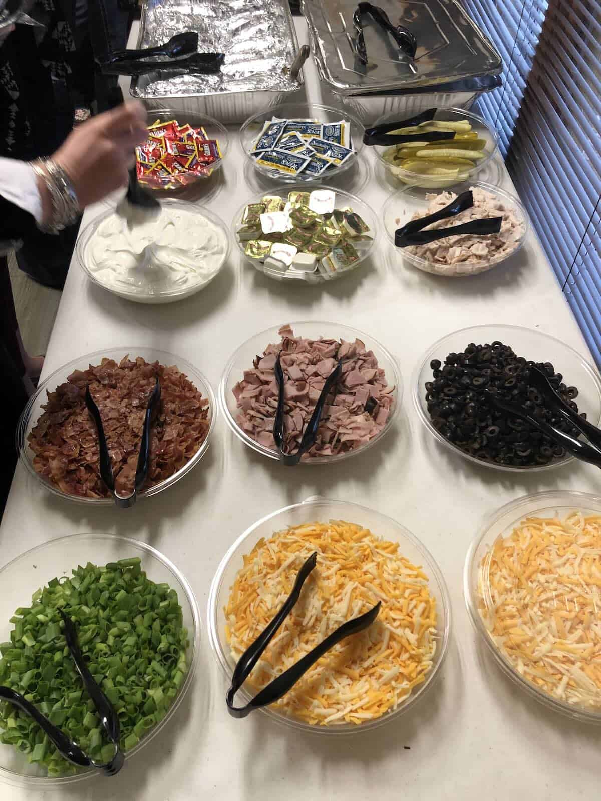 A table full of potato toppings like shredded cheese, sour cream, diced ham, green onions, black olives and butter.