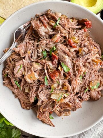 A bowl full of shredded beef with a fork in it surrounded by avocado and flour tortillas.