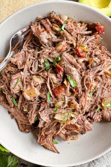 A bowl full of shredded beef with a fork in it surrounded by avocado and flour tortillas.