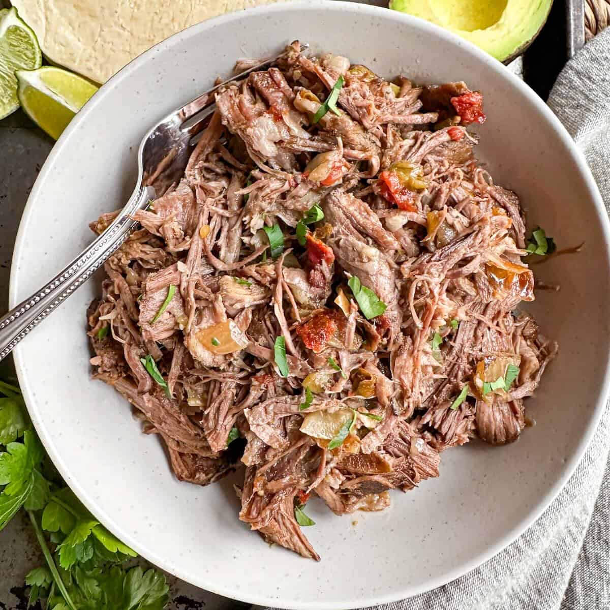 A bowl full of shredded beef with a fork in it surrounded by avocado and flour tortillas.