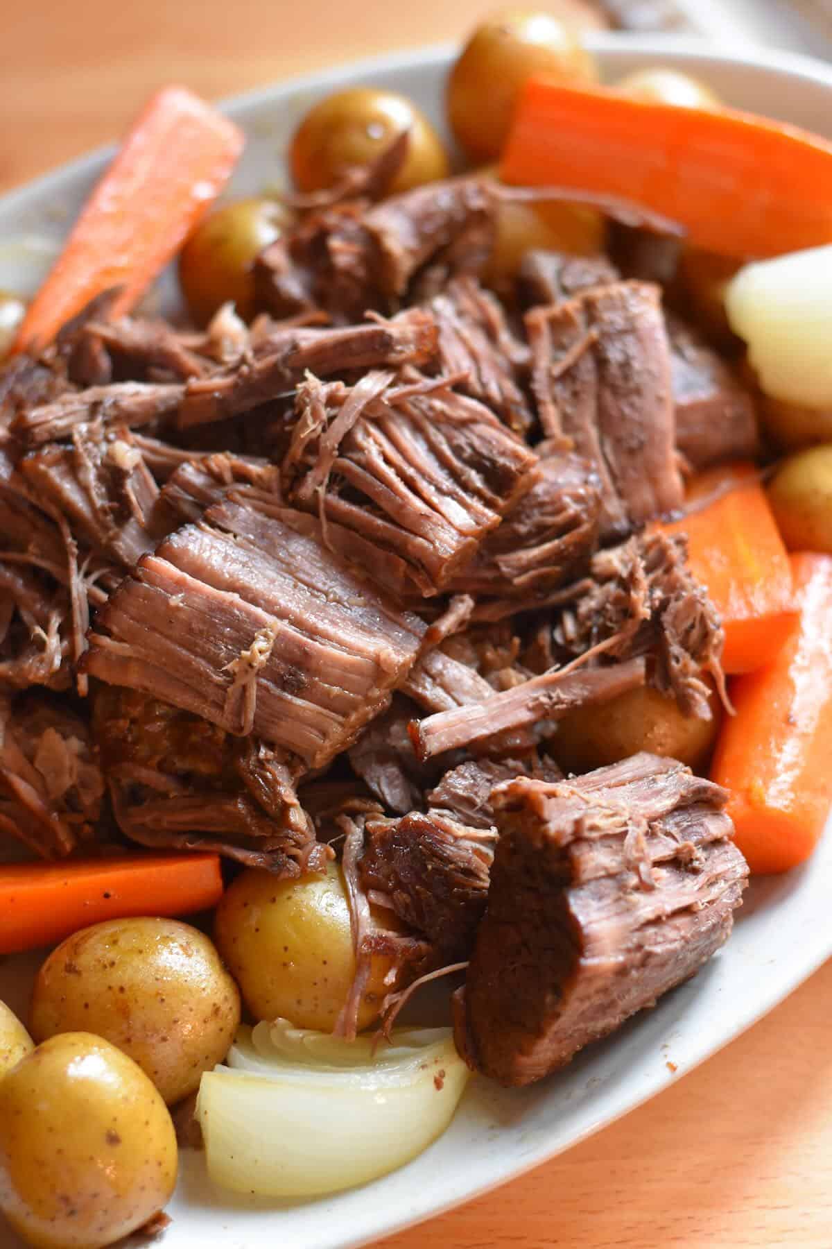 Tender shredded beef heaped on a platter with cooked onions, potatoes and carrots, drizzled with juices.