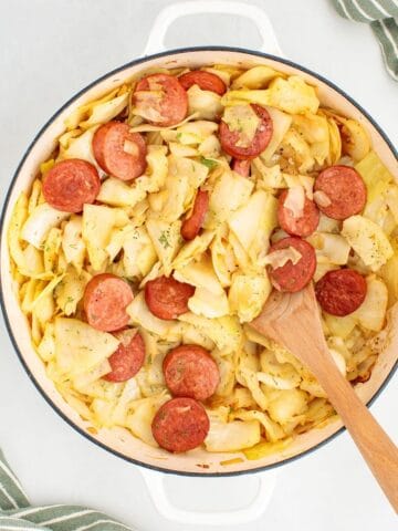 A cooked skillet dinner made with sliced kielbasa and tender cabbage, with a wooden spoon for serving.