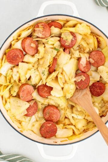 A cooked skillet dinner made with sliced kielbasa and tender cabbage, with a wooden spoon for serving.