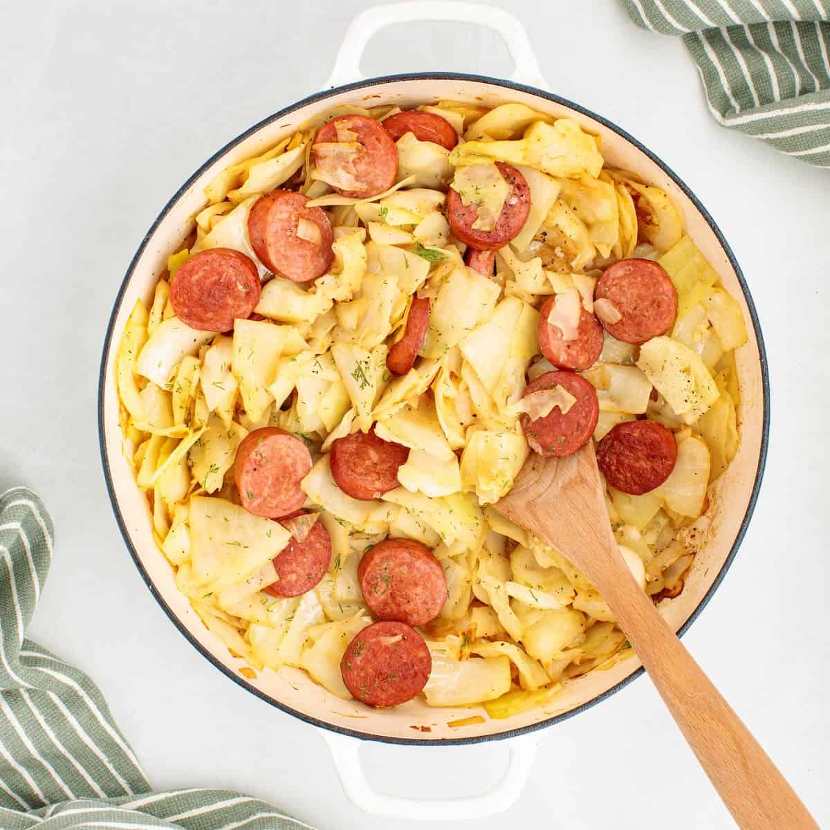 A cooked skillet dinner made with sliced kielbasa and tender cabbage, with a wooden spoon for serving.