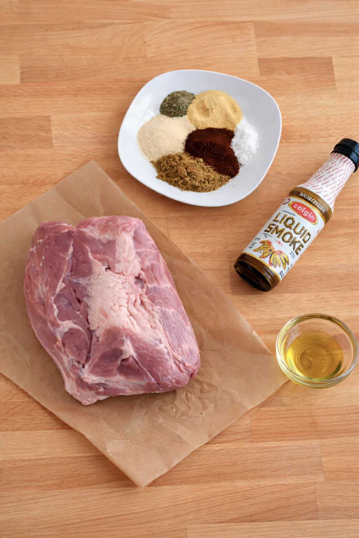 Air fryer pulled pork ingredients including pork shoulder, spice rub, olive oil, and liquid smoke on a butcher block countertop.