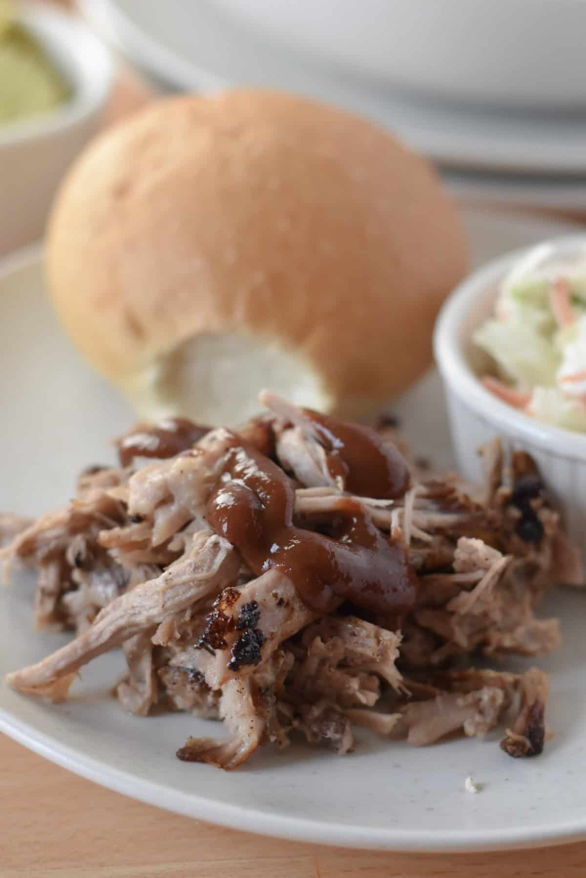 Shredded air fryer pulled pork topped with barbecue sauce on a plate to serve with a roll and coleslaw.