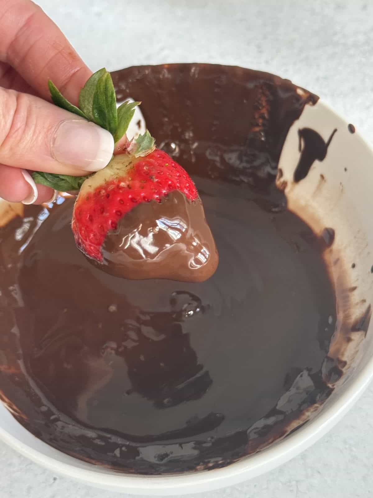 Strawberry being dipped into melted chocolate while held by the green stem.