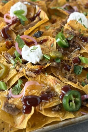 Close-up of leftover BBQ pulled pork nachos with melted cheese, barbecue sauce, pickled red onions, jalapeños, and sour cream.