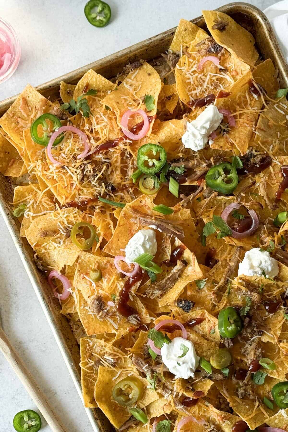 Sheet pan of leftover BBQ pulled pork nachos topped with melted cheese, pickled red onions, jalapeños, and barbecue sauce.
