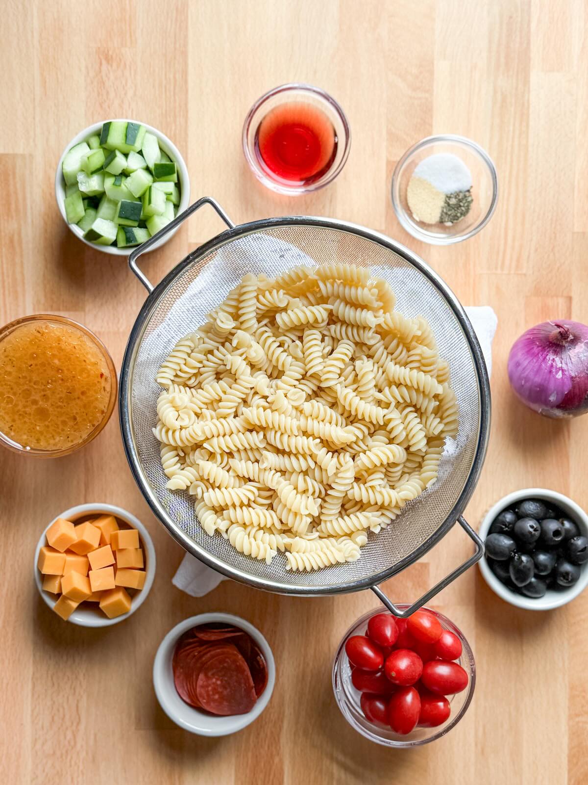 Ingredients for Italian pasta salad including rotini pasta, tomatoes, cucumber, cheese, and Italian dressing.