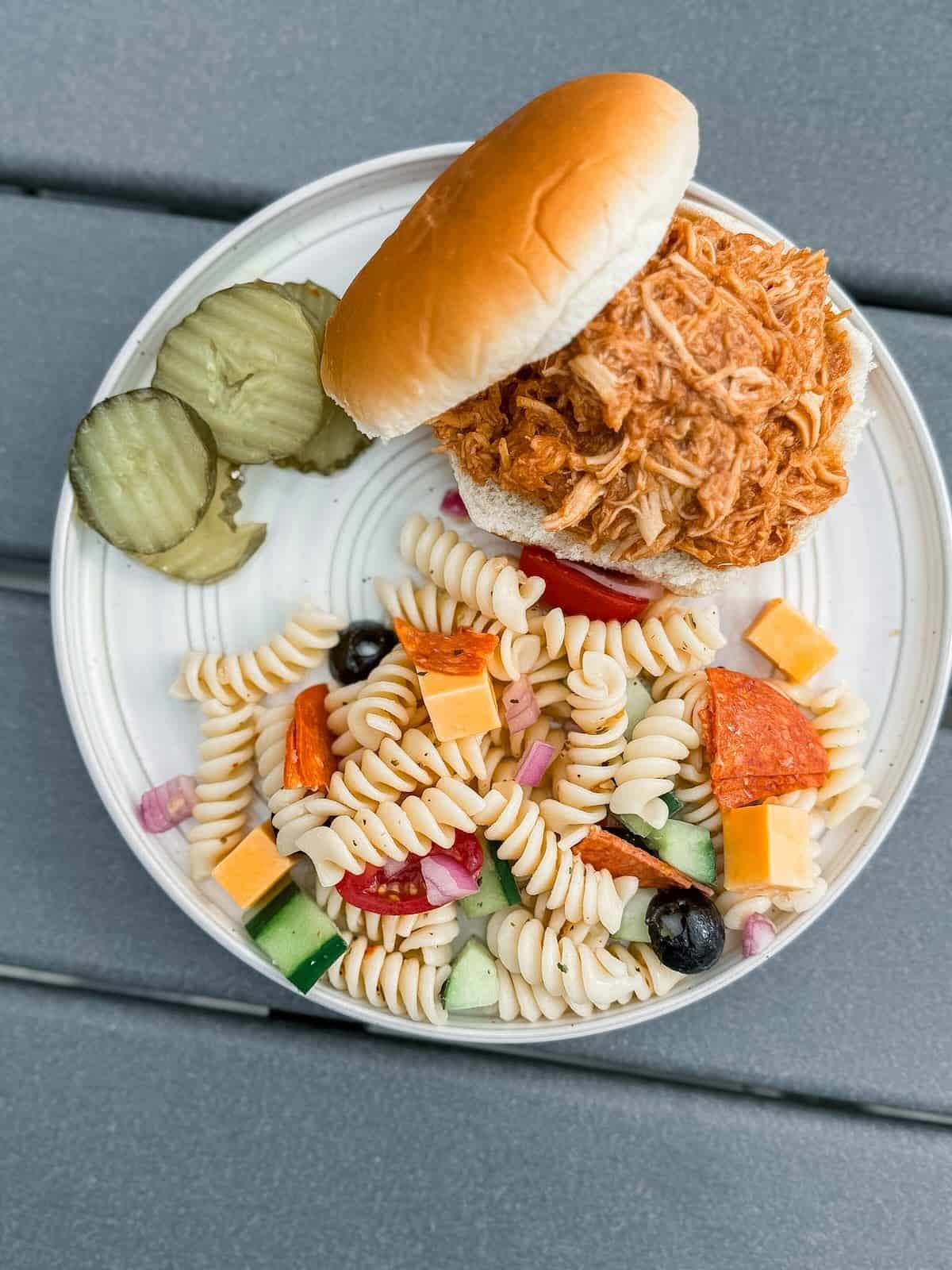 A plated meal of shredded bbq chicken on a bun with Italian pasta salad and pickle slices.