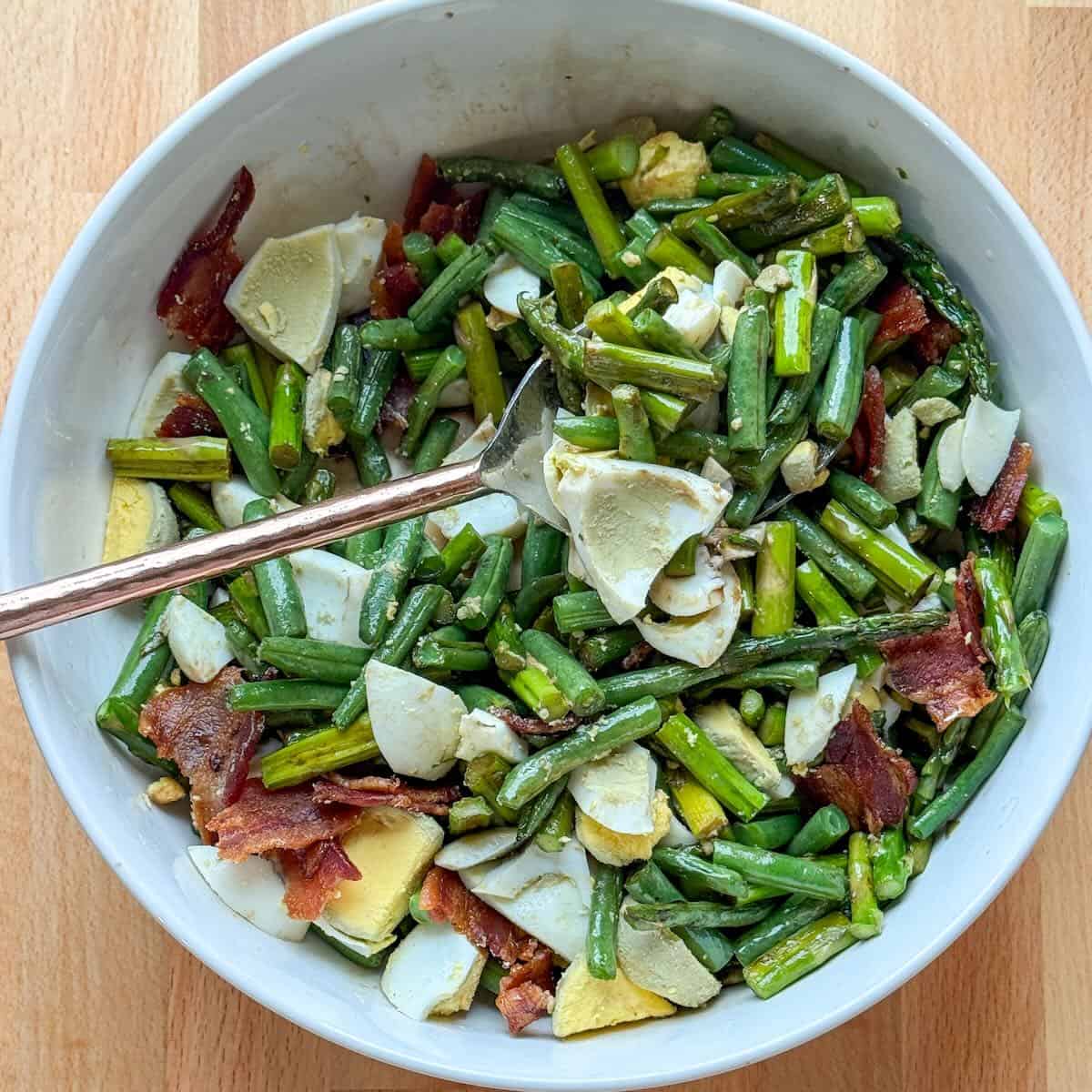 Roasted asparagus and green beans topped with bacon and hard boiled eggs in a serving bowl.