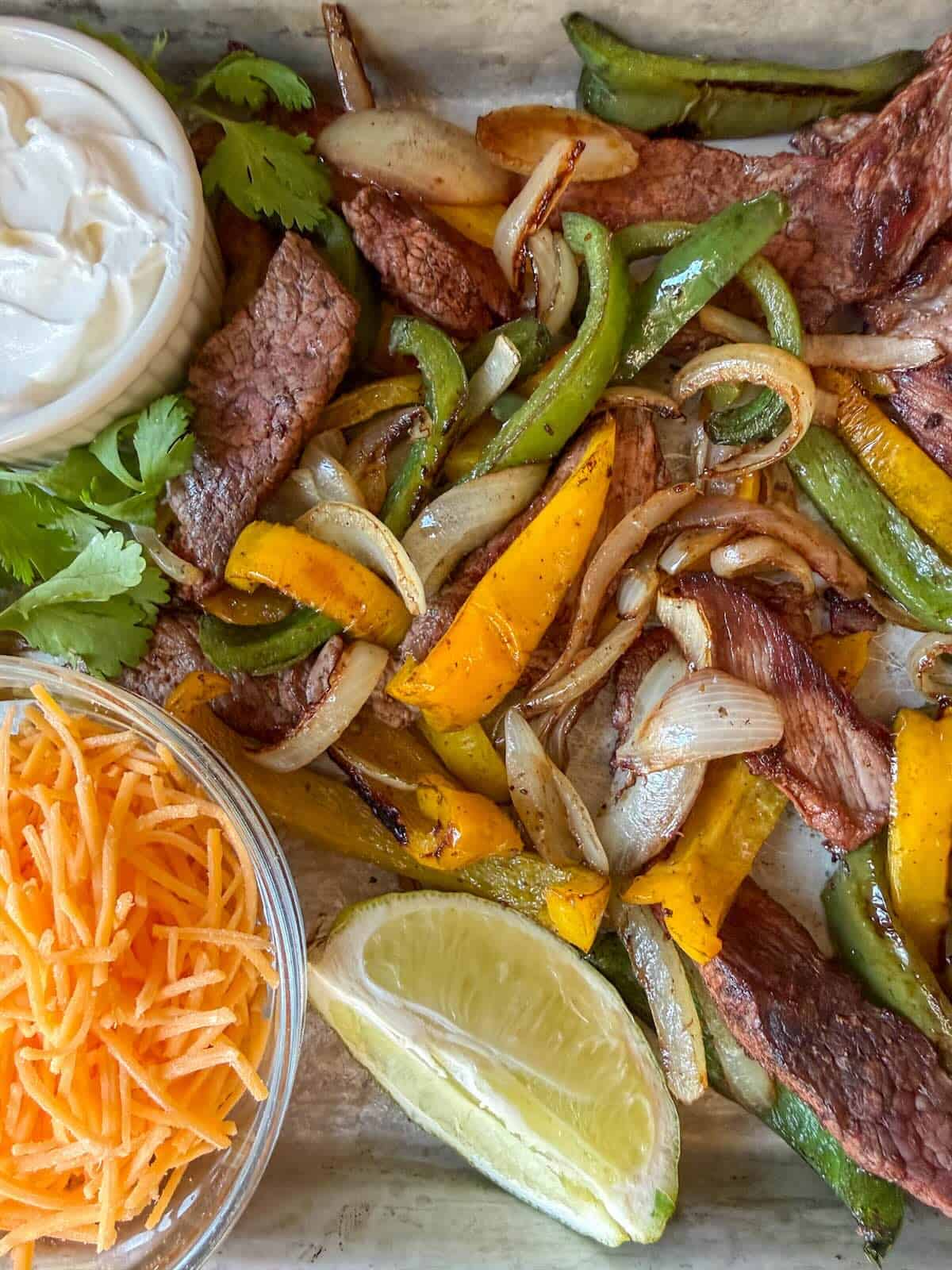 Fajitas made in the oven, ready to serve with cheese, lime wedges, cilantro and sour cream.