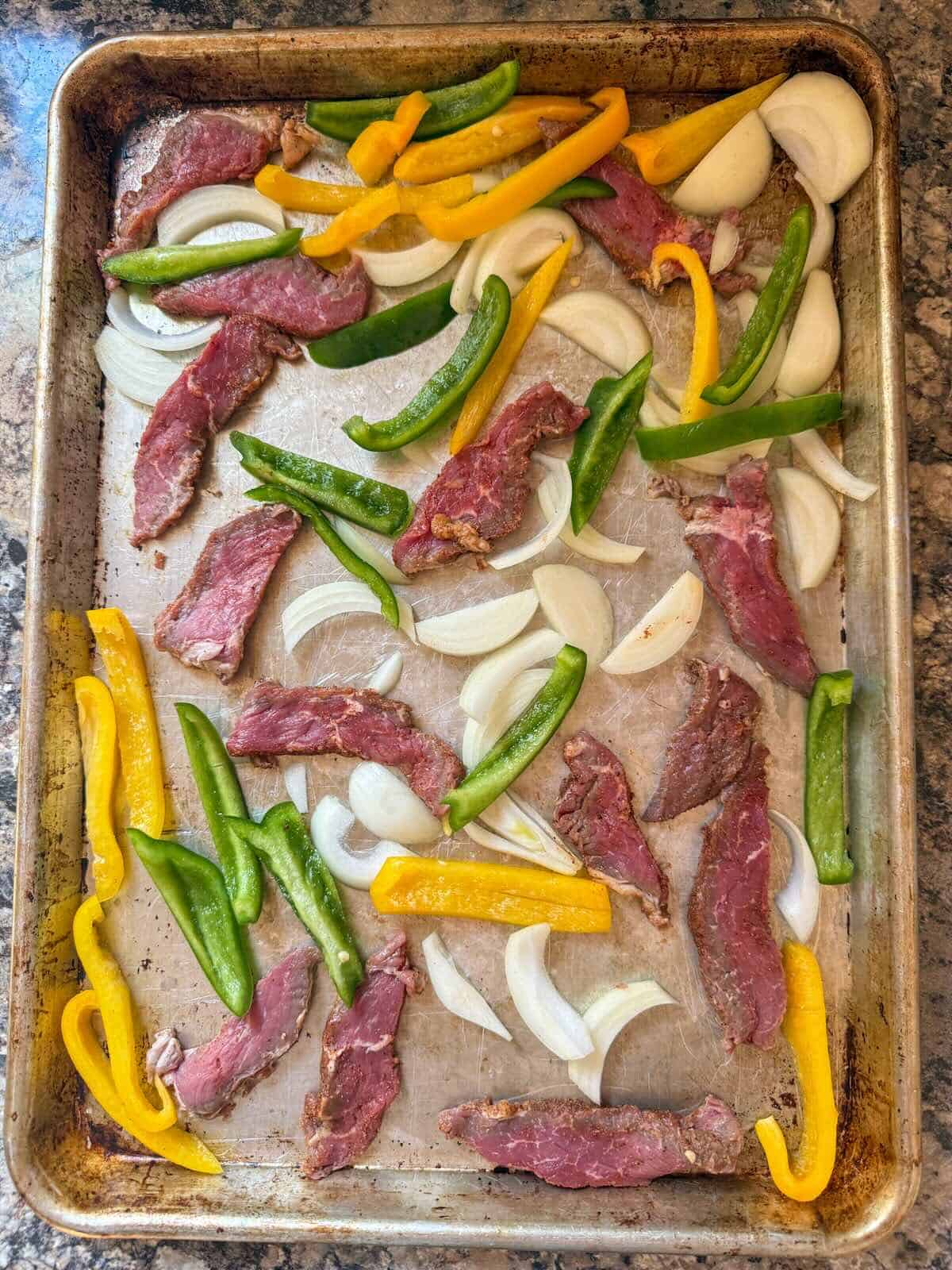 Sliced steak, bell peppers and onions on a sheet pan before cooking into oven baked fajitas.