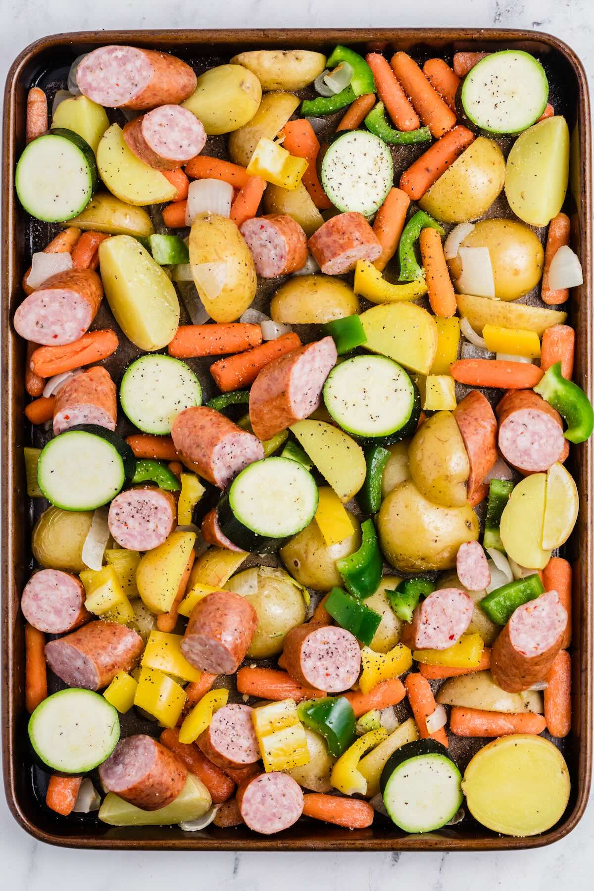 A sheet pan dinner filled with kielbasa slices, zucchini, carrots, onion, potatoes and bell peppers, ready to finish roasting in the oven.