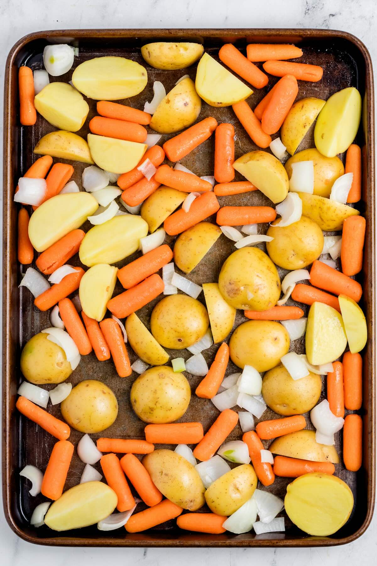 A sheet pan filled with cut potatoes, baby carrots and chopped onion before roasting.