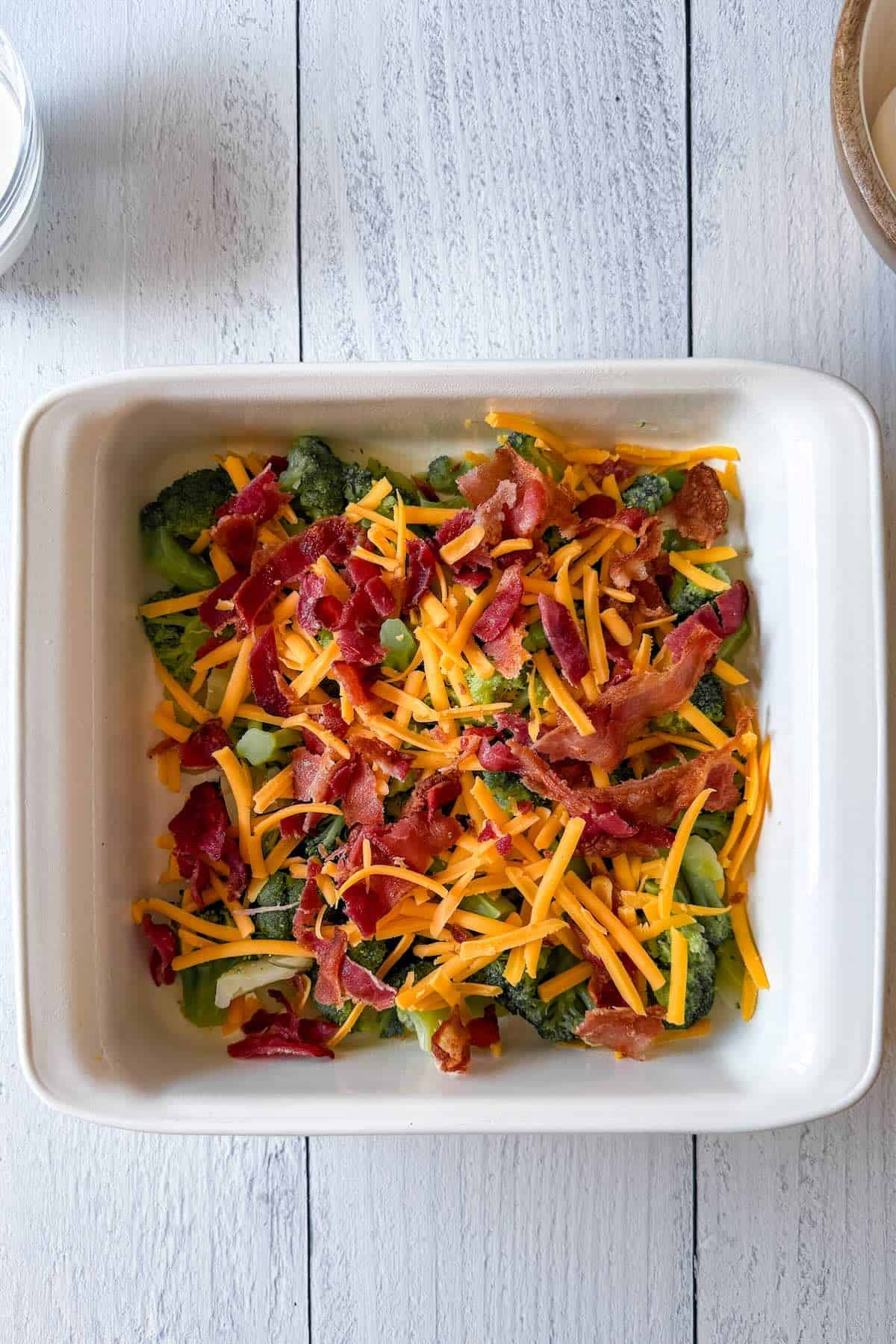 Broccoli, bacon, and shredded cheese layered in a baking dish for breakfast casserole.