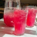 Two glasses of refreshing blueberry lemonade with a pitcher of more to be poured.