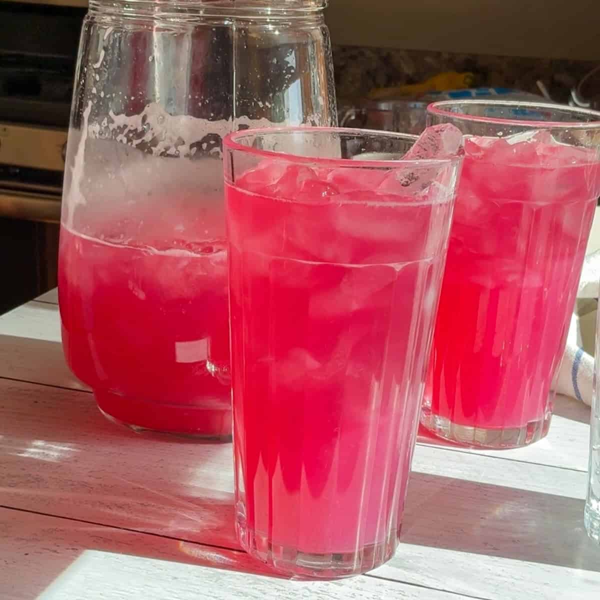 Two glasses of refreshing blueberry lemonade with a pitcher of more to be poured.