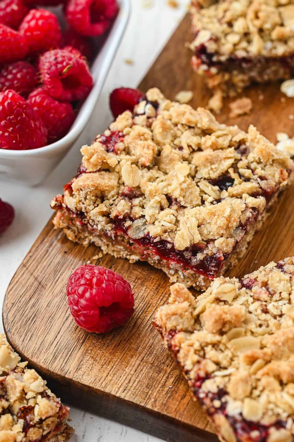 Raspberry oatmeal bars with a buttery oat crumble topping and raspberry jam filling cut into squares on a cutting board.