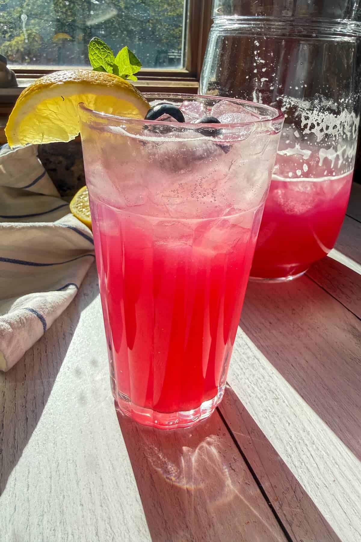 A glass of sparkling blueberry lemonade garnished with mint, fresh blueberries and lemon wedges.