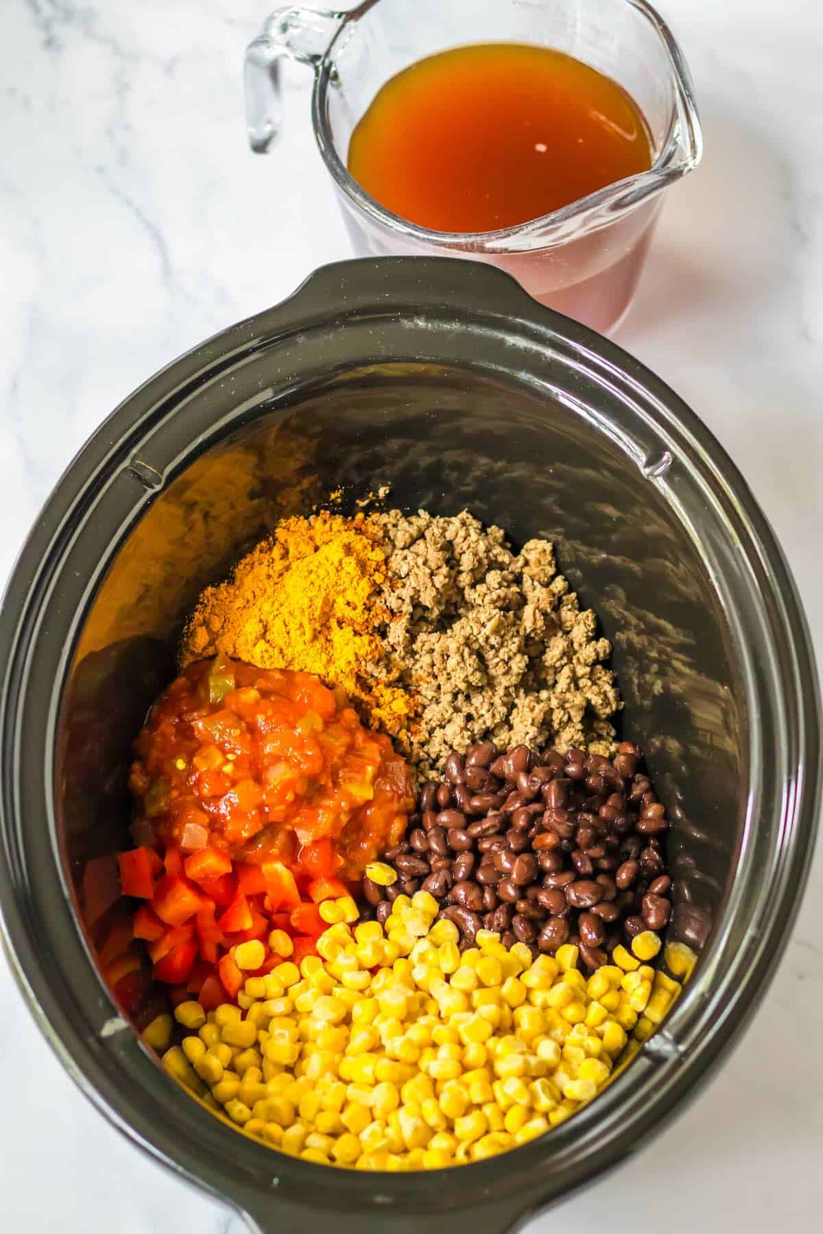 Beef broth being added to ground beef, salsa, corn, black beans, peppers in a slow cooker for taco pasta.