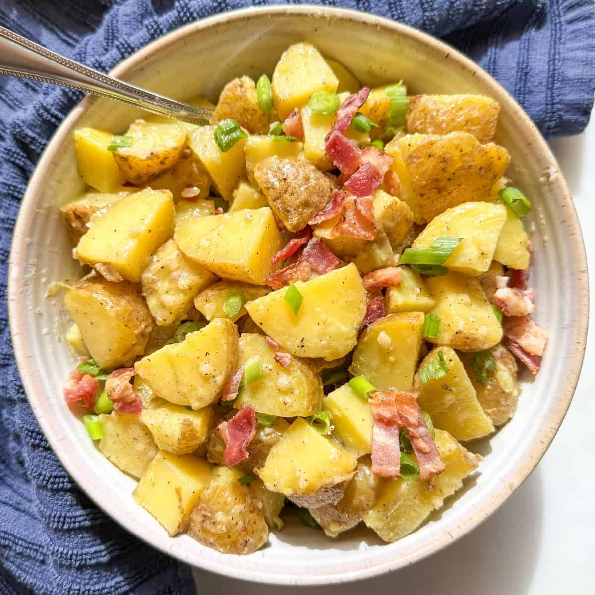 Rustic bowl of potato salad without mayo garnished with crispy bacon and sliced green onions.