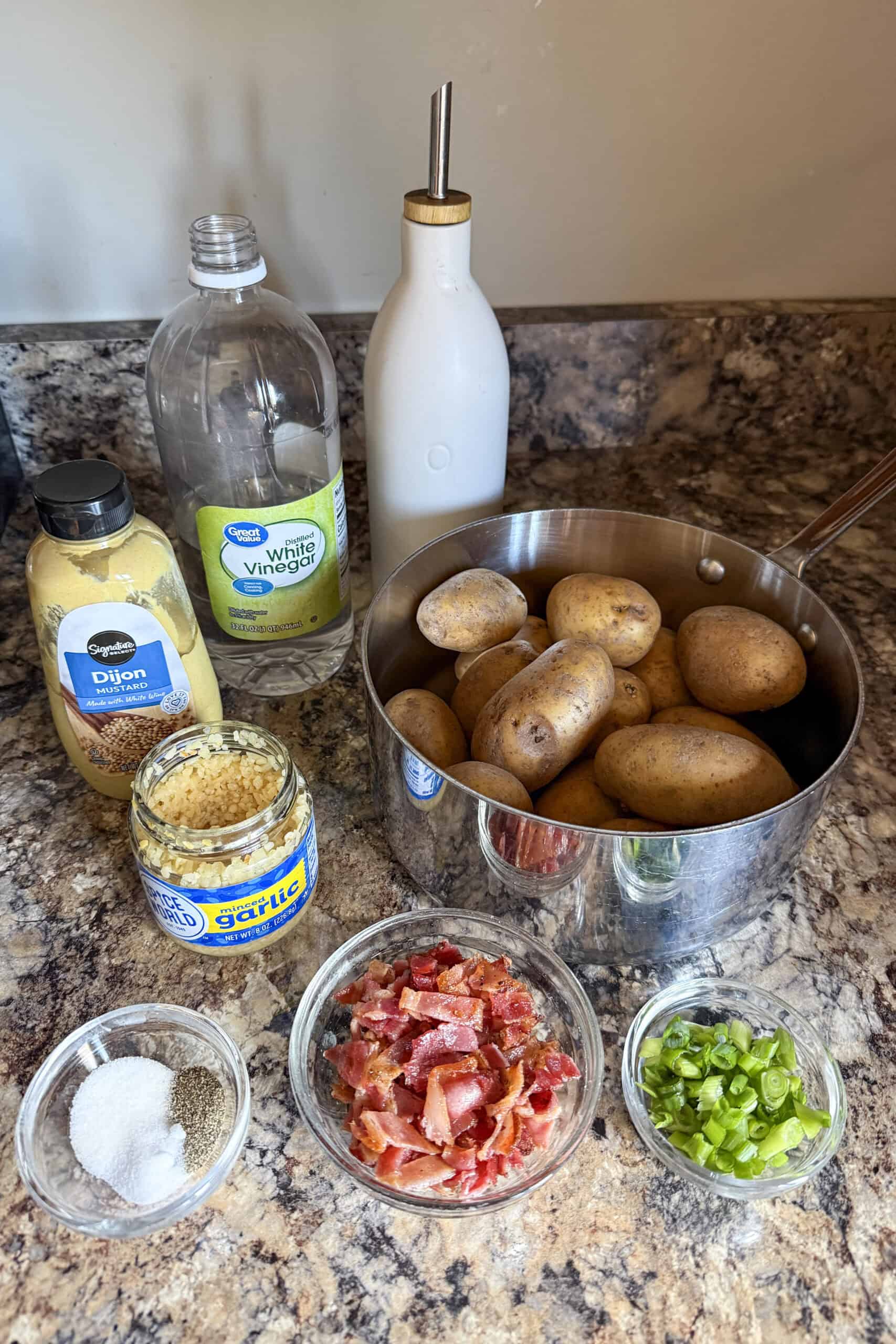 Ingredients for no mayo potato salad including Yukon gold potatoes, bacon, green onions, olive oil, vinegar, garlic, and Dijon mustard arranged on a kitchen counter.