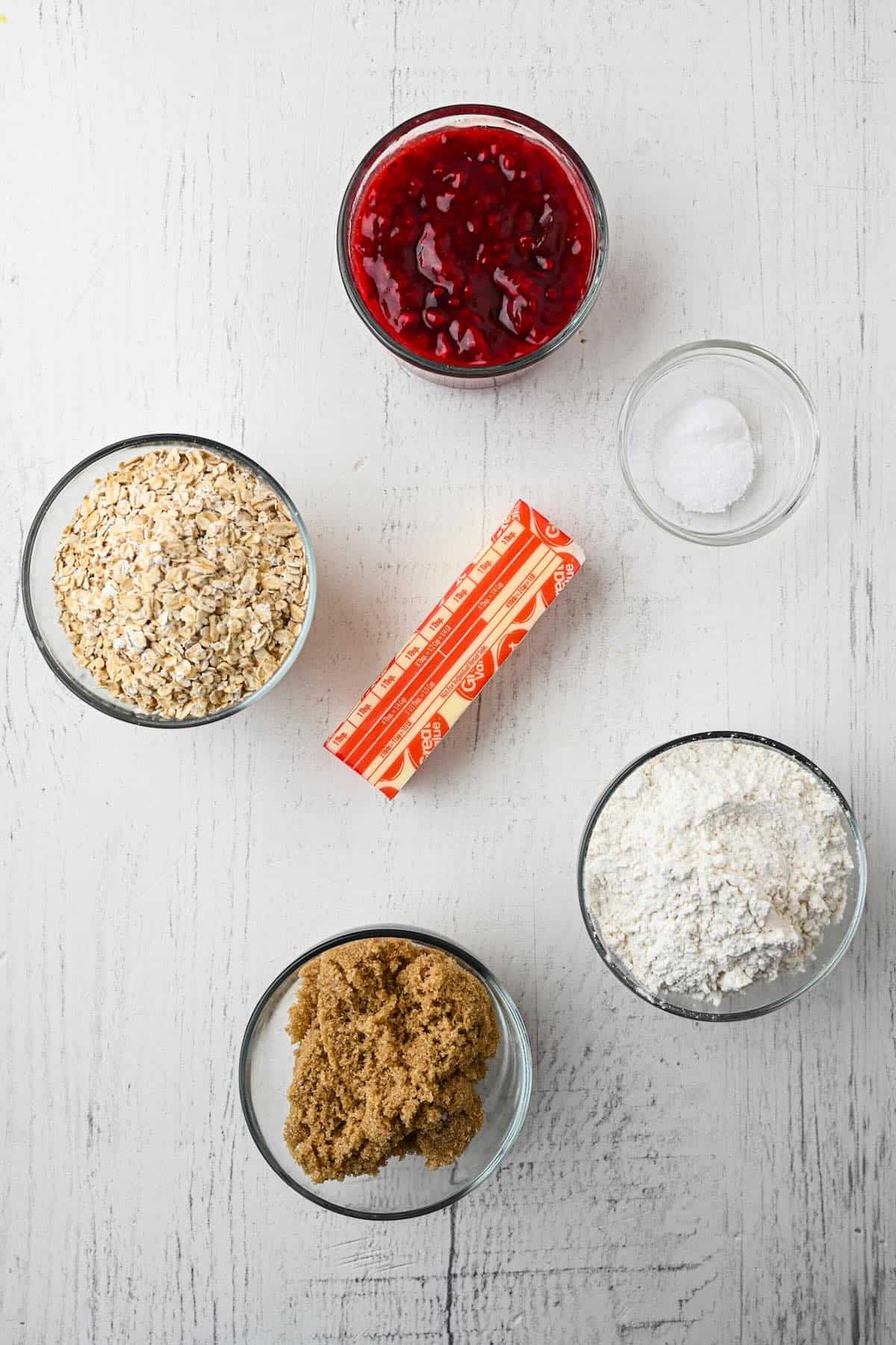 Ingredients for raspberry oatmeal bars including oats, flour, brown sugar, butter, and raspberry jam.