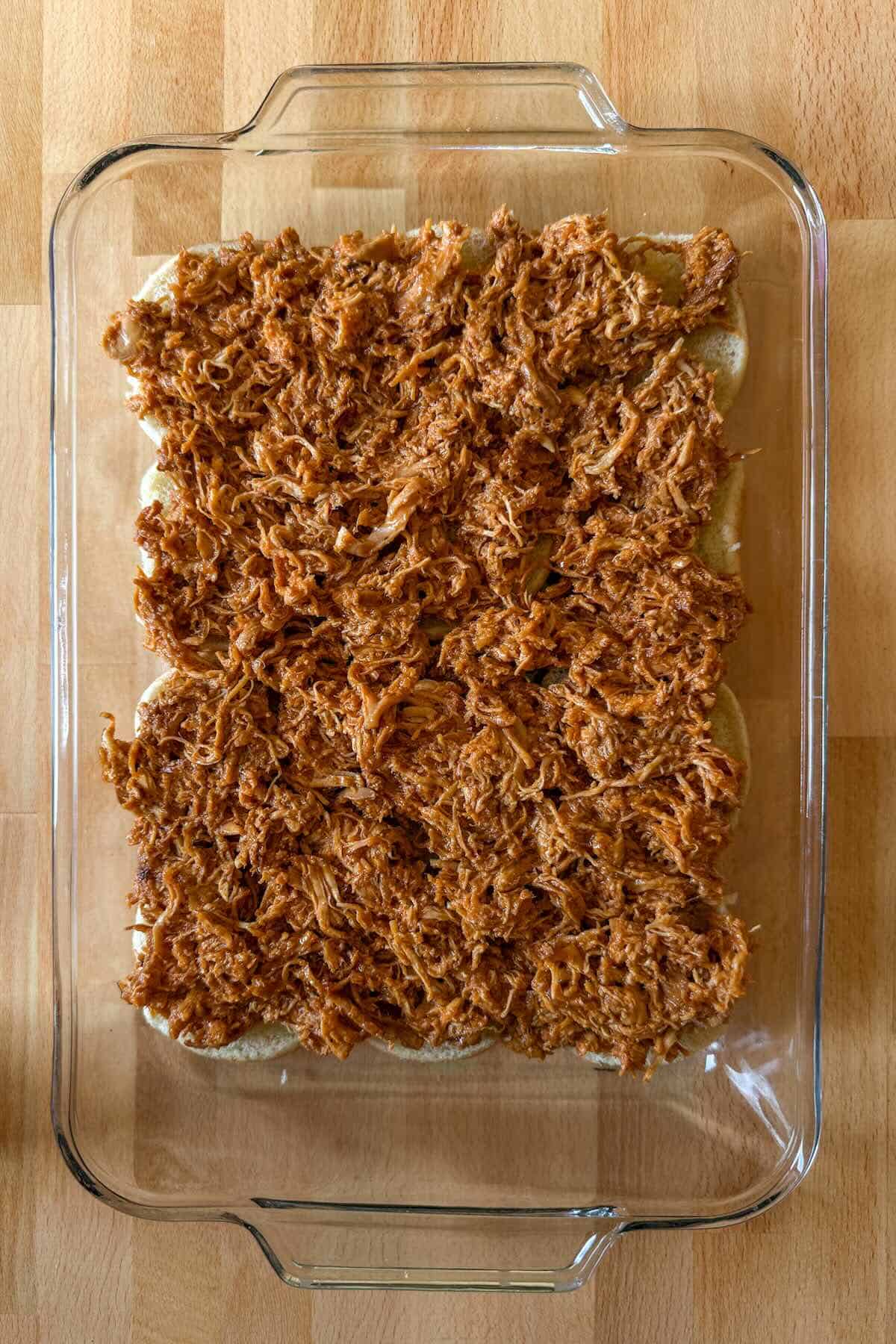 Shredded bbq chicken spread over buns in a glass casserole dish.