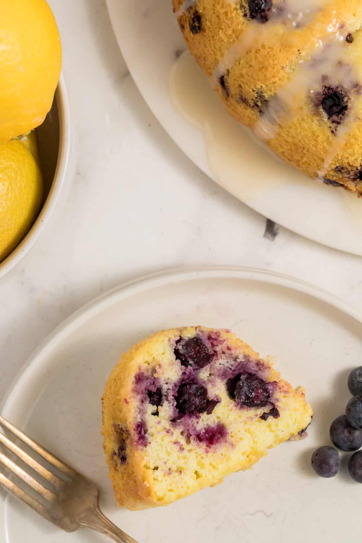 Slice of lemon blueberry bundt cake served on a white plate with the bundt cake in the background.