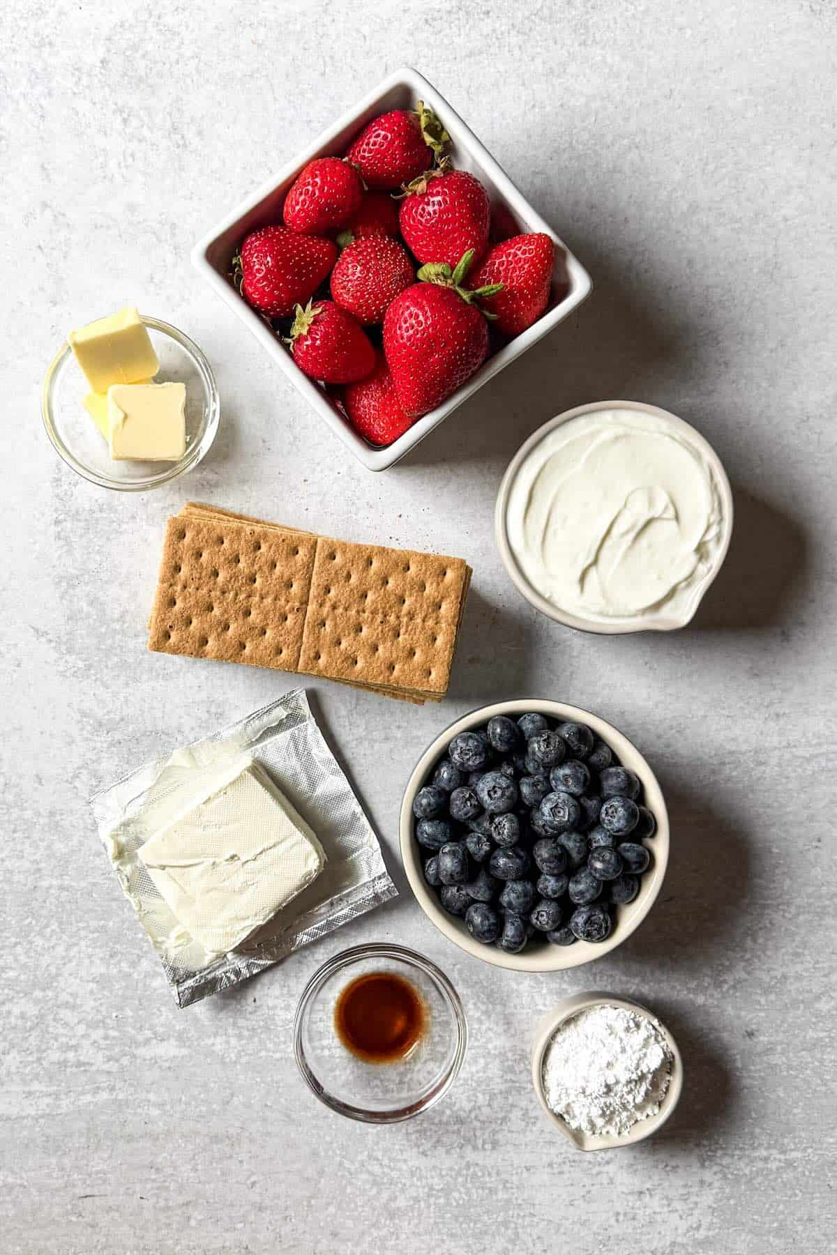 Ingredients for no bake cheesecake parfaits including cream cheese, Greek yogurt, graham cracker crumbs, strawberries, and blueberries.