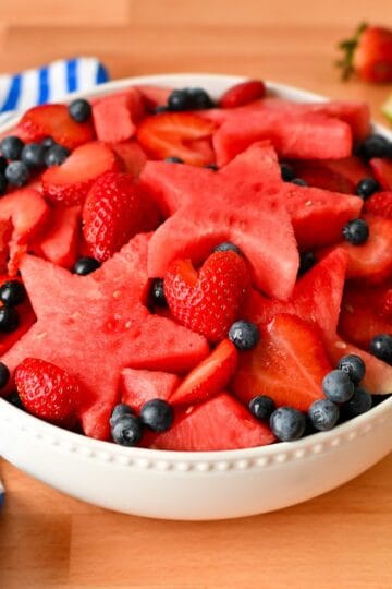 Red white and blue fruit salad with watermelon stars, sliced strawberries, and blueberries in a white bowl.