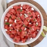 A cutting board and bowl with a watermelon feta salad and lime wedges, ready to serve.