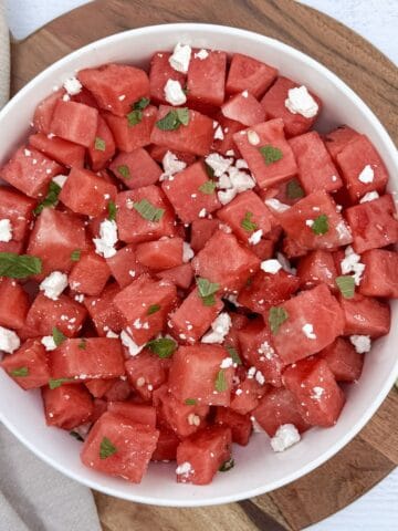 A cutting board and bowl with a watermelon feta salad and lime wedges, ready to serve.