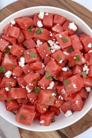 A cutting board and bowl with a watermelon feta salad and lime wedges, ready to serve.