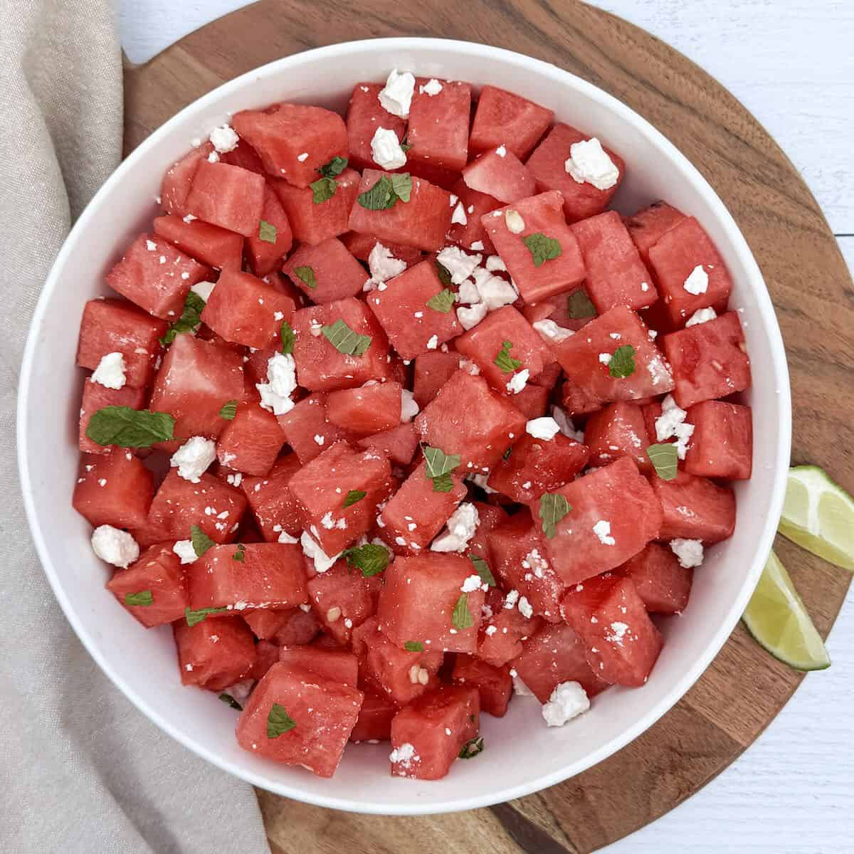 A cutting board and bowl with a watermelon feta salad and lime wedges, ready to serve.