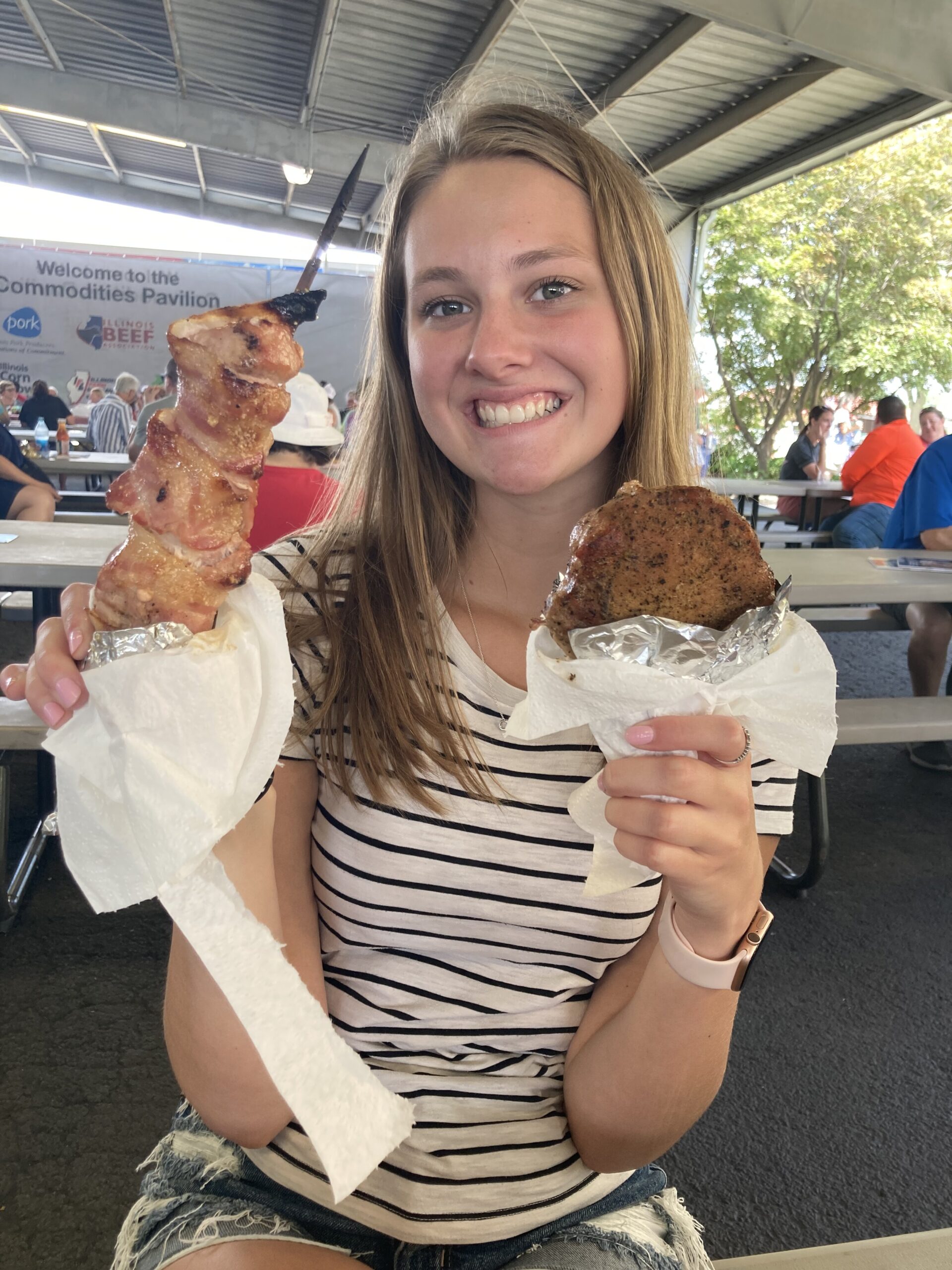 Ava holding a pork burger and a bacon wrapped pork kabob at the fair.