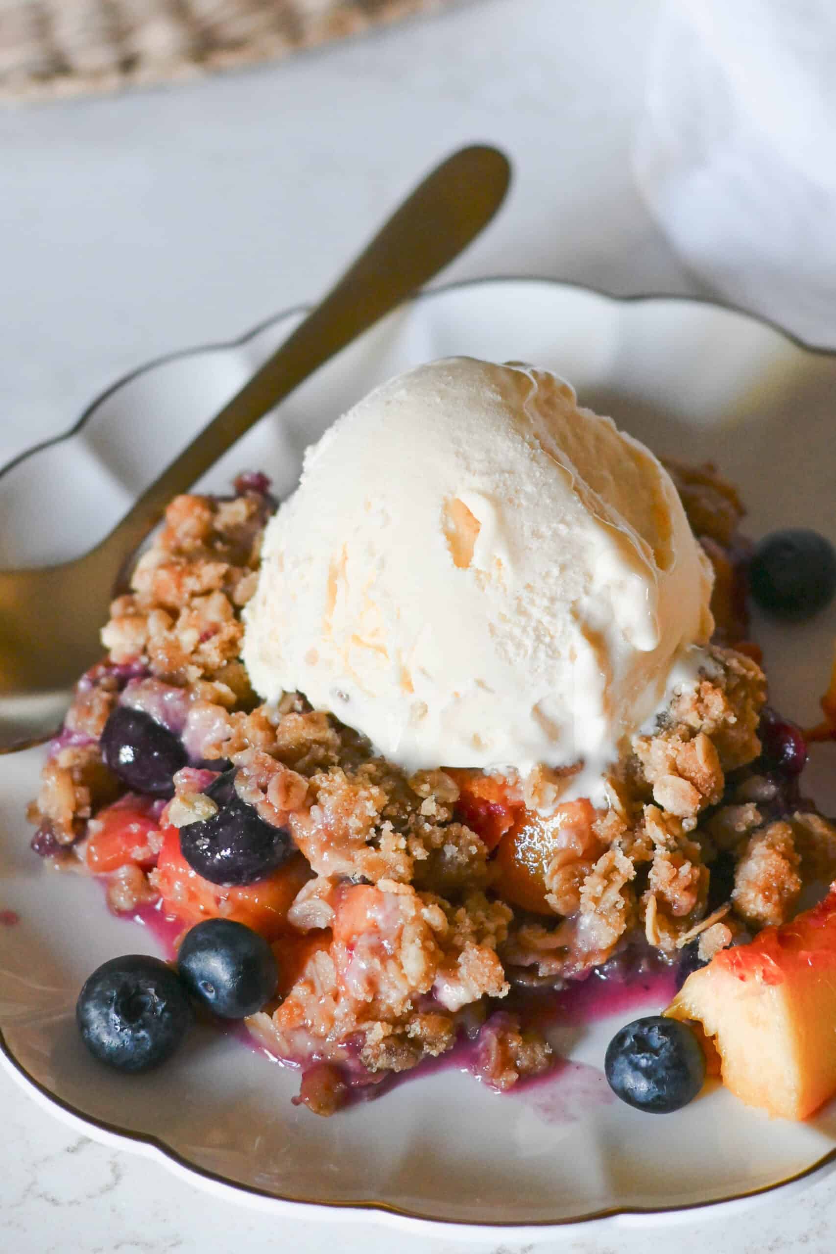 A peach and blueberry crisp topped with a scoop of vanilla ice cream.