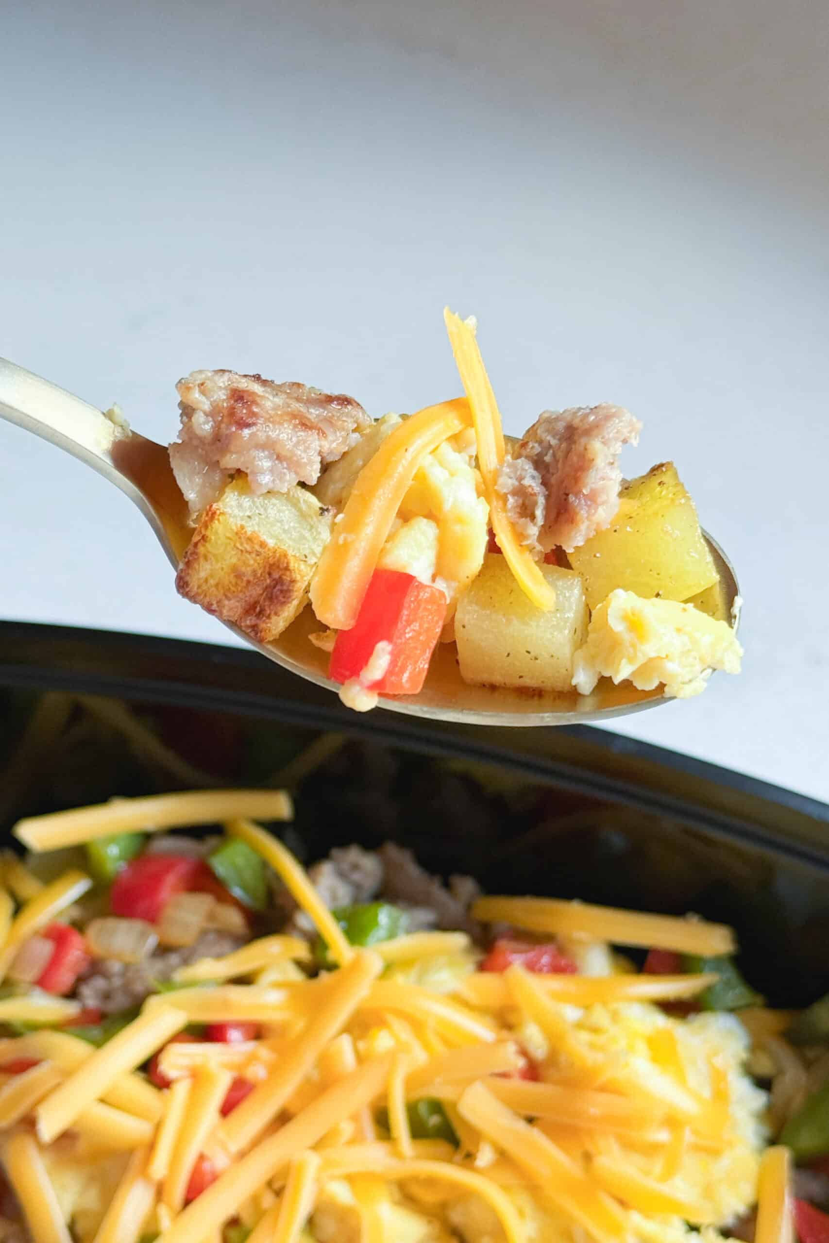 A spoon loaded with roasted potato, shredded cheee, eggs, sausage and bell peppers.