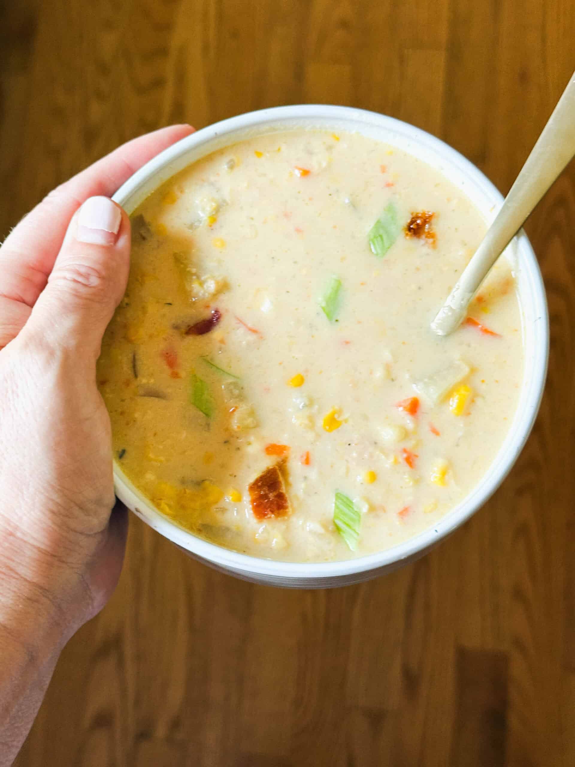 A hand holding a bowl of steamy corn chowder garnished with bacon.