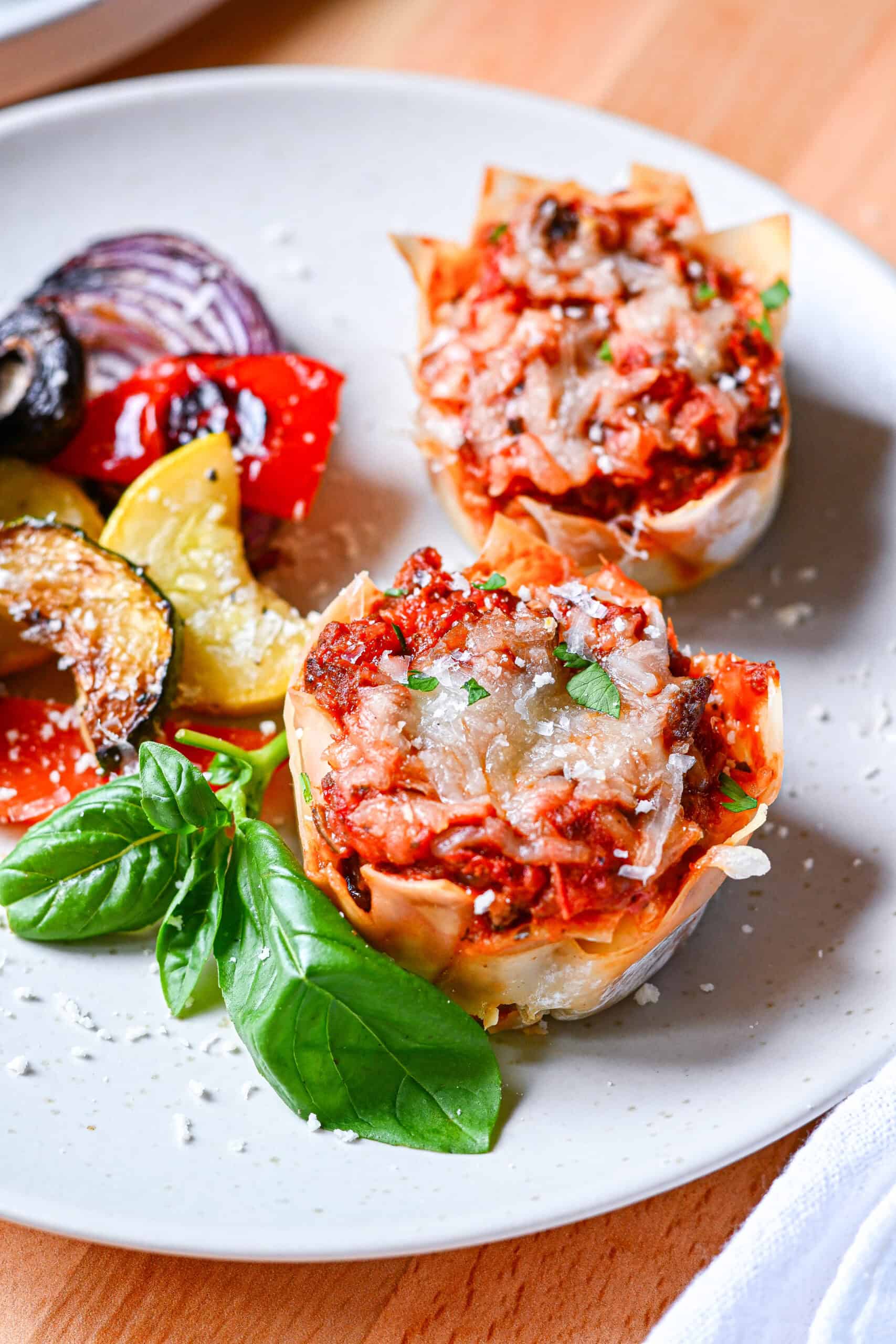 Two baked lasagna cups with melted cheese on a plate with grilled vegetables and fresh basil.