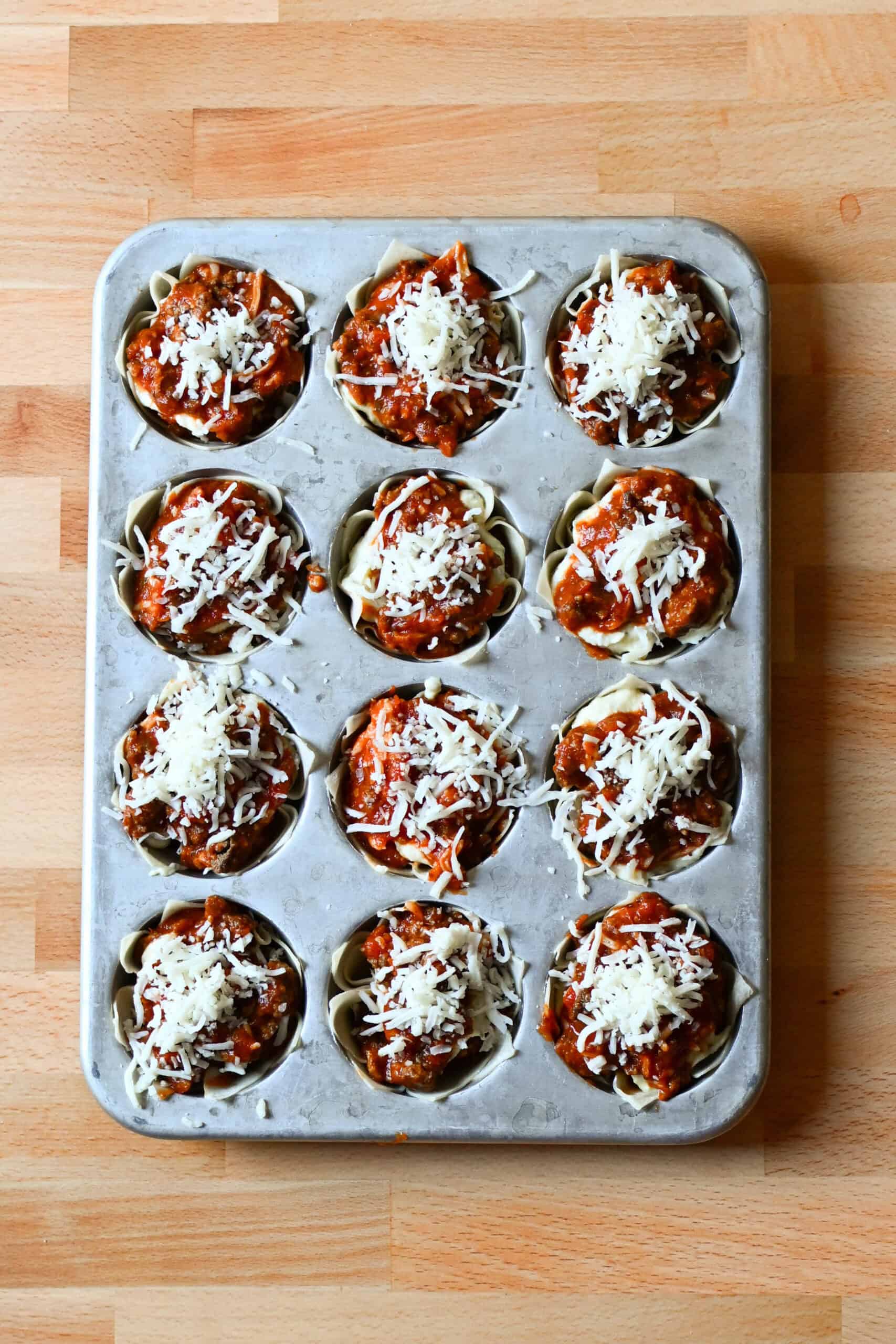 Unbaked lasagna cups in a muffin tin topped with meat sauce and shredded mozzarella.